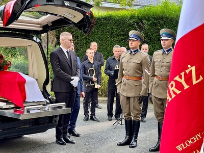 Na pierwszym planie sztandar, obok pojazd z trumną, przy pojeździe funkcjonariusze Na pierwszym planie sztandar, obok pojazd z trumną, przy pojeździe funkcjonariusze